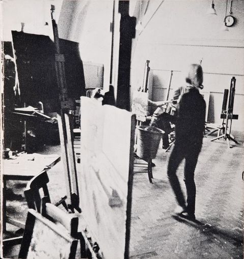 Black and white photograph. Two people in a studio space with paintings on easels. One person is blurry as they walk across the space.